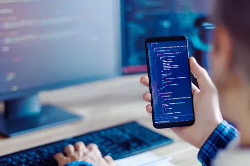La main d'un travailleur indépendant tient un smartphone avec un code de programme. Le développeur écrit un logiciel sur plusieurs écrans d'ordinateur dans son bureau à domicile. Appareils de travail. Concept de lieu de travail pour geek.